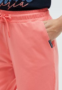 Korallfarbene Joggerhose mit elastischem Bund und verstellbarem Kordelzug, ausgestattet mit Seitentaschen und einem kleinen Markenetikett auf der Tasche.