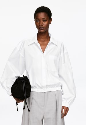 Kobieta ubrana w luźną białą koszulę na guziki i jasnoszare spodnie, trzymająca czarną torbę ze ściągaczem na jednolitym białym tle.