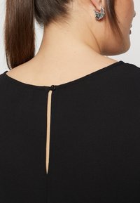 Crna bluzica sa zatvaranjem na dugmad na leđima i dizajnom rezanog ključanika. Materijal izgleda teksturirano s glatkom završnicom.