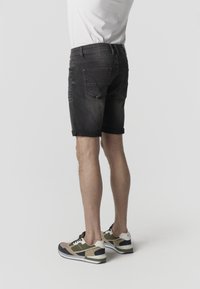 Mees, kes kannab rullitud musta teksapükstega ja beeže, musta ning rohelist värvi vabaajajalatseid, seisab ühevärvilise tausta ees.