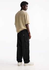 T-shirt beige oversize abbinato a pantaloni cargo neri con tasche e etichetta sul retro. Il modello indossa sneakers bianche.