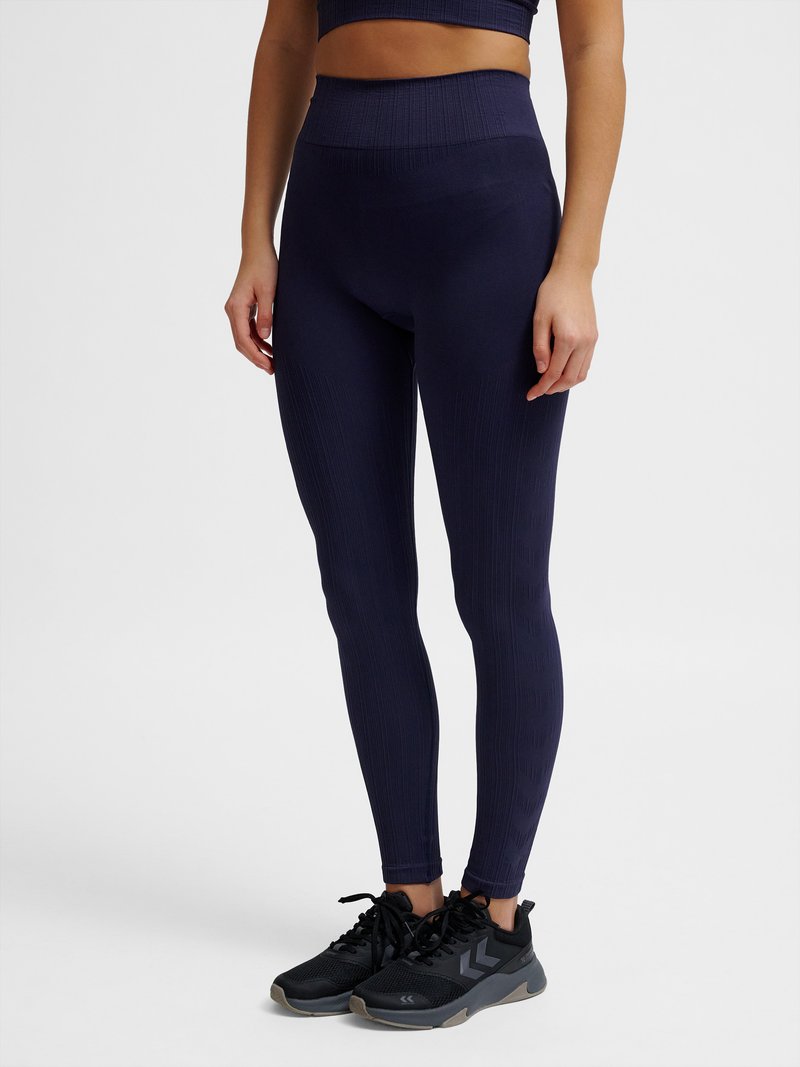 Kobieta nosząca ciemnoniebieskie legginsy z wysokim stanem i czarne buty sportowe stojąca na jednolitym tle.
