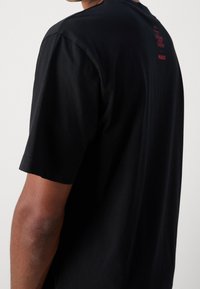 Musta puuvillainen T-paita, jossa on lyhyet hihat, pyöreä kaula-aukko ja huomaamaton punainen tekstidetaljointi selkäpuolella, mikä viittaa brändäykseen.