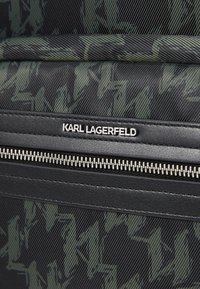 Black textile bag featuring a green geometric camo pattern, leather accents, a zippered closure, and a metallic logo label.