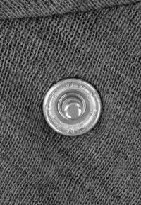 Metallknopf auf grauem Strickstoff, mit runder Form, glänzender Oberfläche und eingravierten Markierungen rund um das Druckknopfloch.