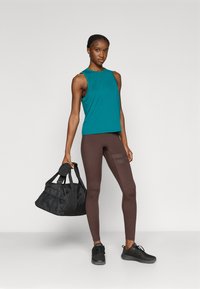 Μπλε-πράσινο αμάνικο τοπ, καφέ κολάν με δίχτυ στα πλαϊνά και μαύρα αθλητικά παπούτσια. Το μοντέλο κρατά μια μαύρη γυμναστηριακή τσάντα με δύο χερούλια.