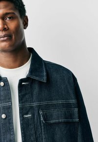 Donker denimjacket met een knoopsluiting, een borstzak en witte stiksels. Gedragen over een wit shirt met een klassieke kraag.