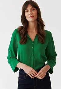 Bluză verde, cu mâneci lungi și guler, având un fațadă cu nasturi și detalii la mansete. Textură netedă, material ușor și croială lejeră.
