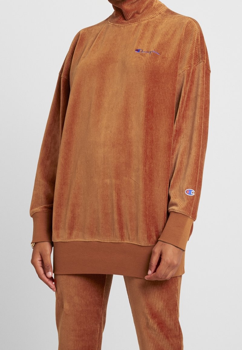 Tunic in corduroy marrone con collo alto, texture a costine e maniche allungate. Presenta un piccolo logo ricamato sul petto.