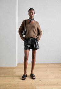 Brun strikket sweater, sorte lædershorts med bælte, sorte chunky sandaler og et sort-hvidt tørklæde, sat mod en neutral baggrund.
