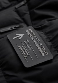 Schwarze Steppjacke mit matter Textur, versehen mit einem rechteckigen schwarzen Etikett mit erhabener Schrift und Pfeildetails, das am Stoff angebracht ist.