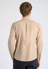 Camicia beige a maniche lunghe con tessuto testurizzato, orlo arrotondato e polsini con bottoni. Il retro presenta una piega centrale per un maggiore movimento.