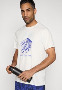 T-shirt de sport blanc avec un motif montagne bleu, manches courtes, et logo sur la manche ; personne tenant une bouteille d'eau noire.