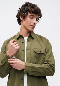 Olíva zöld ing texturált anyagból, gombos kialakítással, foltzsebbel és feltűrt ujjakkal. Fehér trikó alatta.