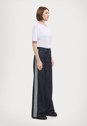 Jeans in denim a gamba larga con un lavaggio scuro, caratterizzati da una striscia laterale grigio chiaro. Abbinati a una t-shirt bianca semplice e calzature nere.