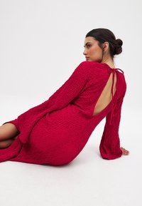 Red textured dress with long sleeves, open back, and bow detail at the neckline. Features a relaxed fit and slight slit at the hem.