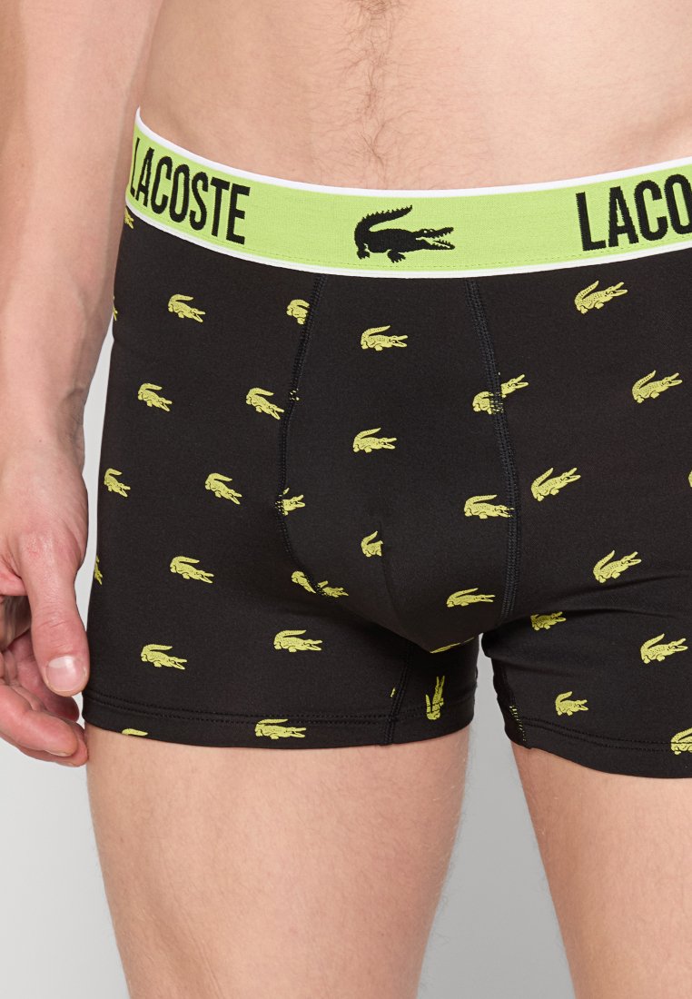Czarne bawełniane bokserki z zielonym elastycznym pasem, na którym widnieje napis "LACOSTE" oraz powtarzające się żółte logo krokodyla.