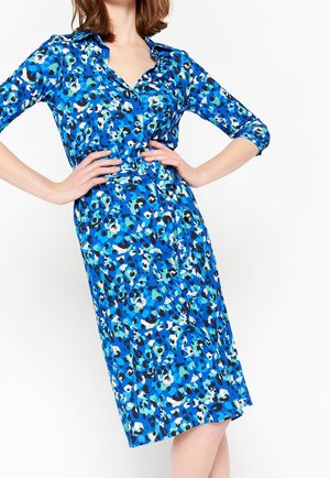 Femme portant une robe bleue à motifs longueur genoux avec manches trois-quarts et ceinture, les mains posées sur sa taille.