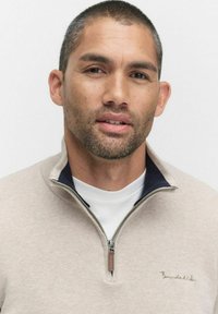 Ljusbeige zip-up tröja med hög krage, med en marinblå accent. Texturerad stickad tyg med en subtil logo broderad på bröstet.