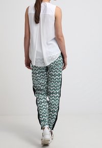 Donna che indossa una blusa senza maniche bianca e pantaloni patterned verdi, neri e bianchi con strisce nere ai lati, mentre si allontana su uno sfondo semplice.