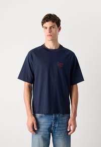 Tamsiai mėlynas trumpomis rankovėmis marškinėliai su raukšlėta apykakle, papuošti raudonu "Calvin Klein" logotipu. Pagaminti iš medvilnės, su laisvu sijonu. Dėvimi su šviesiai mėlynomis džinsomis.