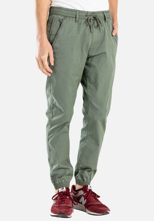 Pantalones jogger de algodón verdes con cintura elástica, cordón, bolsillos laterales y puños ajustados. Llevados con zapatillas deportivas rojas.