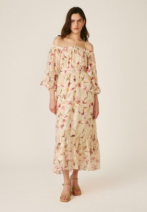 Mujer con un vestido beige floral sin hombros, con mangas 3/4 y dobladillo con volantes, combinado con sandalias con tacón de color tostado, de pie frente a un fondo liso.