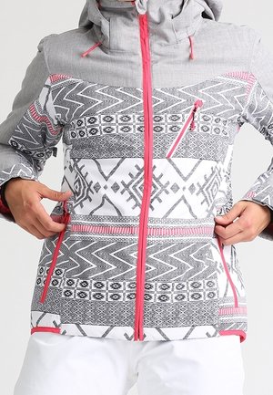 Veste d’hiver avec motifs géométriques gris, noirs et blancs, fermetures éclair roses et une capuche, mains ajustant les poches latérales.