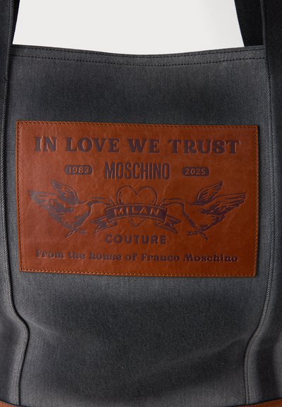 Juodas drobės krepšys su rudos odos pleistru, ant kurio išgraviruota "MEILĖJE PASITIKIME", Moschino logotipas ir paukščių iliustracijos.