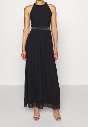 Kobieta w czarnej sukni maxi bez rękawów z dekoltem halter i satynowym paskiem w talii oraz beżowych sandałach na wysokim obcasie.