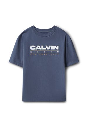 Ung dreng står med hænderne i siden iført blå Calvin Klein t-shirt, løse grå jeans og hvide sneakers på en ensfarvet grå baggrund.