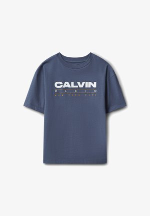 Jeune garçon debout, les mains sur les hanches, portant un t-shirt bleu Calvin Klein, un jean gris ample et des baskets blanches sur un fond gris uni.