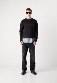 Sweatshirt preto com o logótipo "Boss" em relevo, combinado com uma camisa de colarinho azul claro, jeans escuros e sapatos pretos. Modelo a usar óculos de sol pretos.