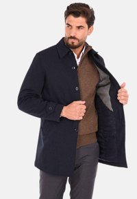 Cappotto blu navy con un design con bottoni, caratterizzato da un colletto classico e una fodera interna grigia, indossato sopra un maglione marrone e pantaloni grigi.