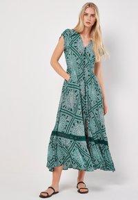 Grünes gemustertes Maxikleid mit V-Ausschnitt, kurzen Ärmeln, Taschen und einem schichten Rock. Der Stoff weist komplexe geometrische Muster auf.
