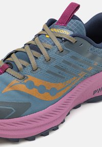Chaussure de trail avec un dessus en mesh bleu, des accents jaunes et une semelle intermédiaire violette. Présente un design texturé et un capuchon renforcé pour plus de durabilité.