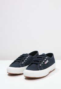 Zapatillas de lona en color azul marino con suelas de goma blanca, puntera redonda y cordones planos. Presentan un pequeño logo en el lateral y un cuerpo de zapato texturizado.
