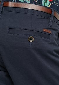 Pantalon bleu marine avec une poche arrière fermée par un bouton en bronze et une étiquette accentuée en cuir synthétique marron. Texture de tissu lisse.