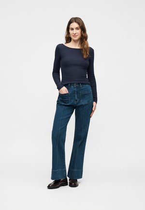 Femme aux longs cheveux bruns portant un haut bleu marine ajusté à manches longues, un jean large taille haute bleu avec de grandes poches avant, et des chaussures noires.