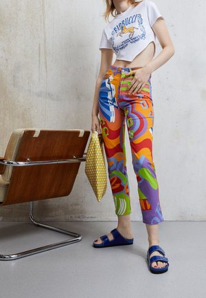 Femme debout portant un pantalon imprimé coloré, un t-shirt court blanc avec un motif de cheval, des sandales bleues, et tenant une pochette jaune à motifs.