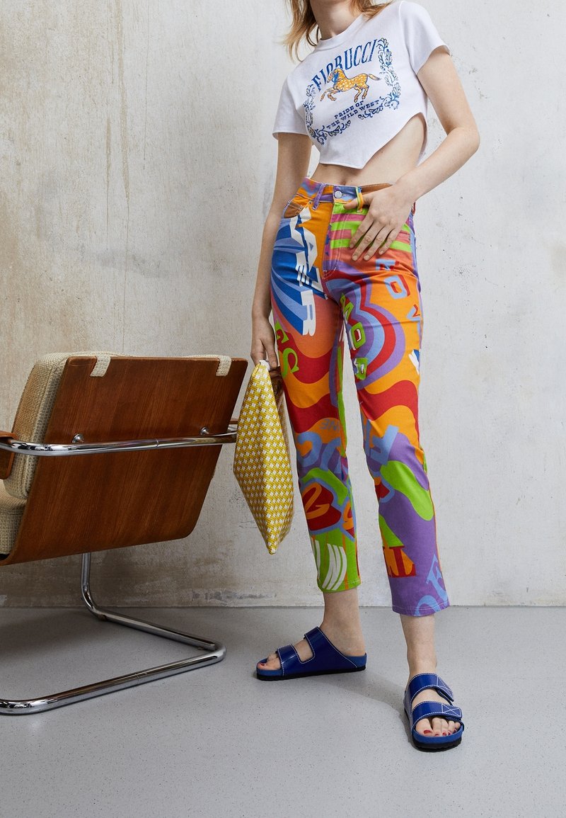 Femme debout portant un pantalon imprimé coloré, un t-shirt court blanc avec un motif de cheval, des sandales bleues, et tenant une pochette jaune à motifs.