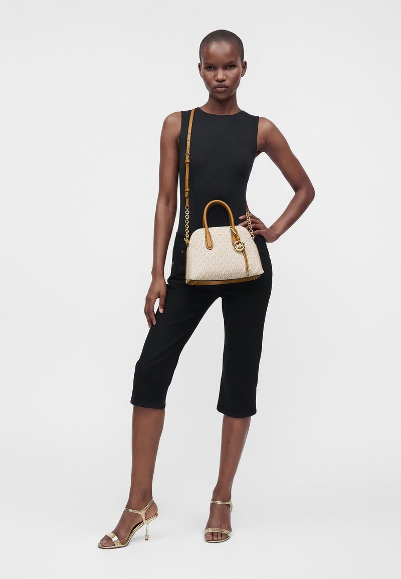 Femme en haut noir sans manches et pantalon court posant avec un sac à main beige et marron et des sandales à talons dorés sur fond blanc.