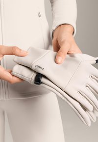 Jasnoszare rękawiczki wykonane z miękkiej tkaniny, z zakładanym designem i czarnym regulowanym paskiem. Na jednej rękawiczce widoczne jest logo "OYSHO".