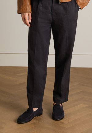 Navy trousers with a slightly textured finish, paired with dark suede loafers. A brown leather jacket complements the outfit. Wood flooring below.