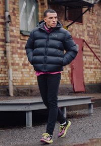 Giacca a vento nera con baffle orizzontali, cappuccio grigio, camicia rosa sotto, pantaloni neri e scarpe da ginnastica multicolori.