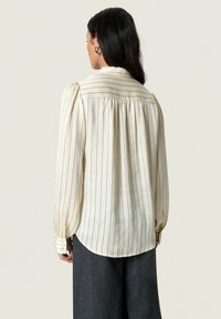 Cream-colored blouse with vertical striped pattern, pleated back, long sleeves, and buttoned cuffs. Fabric has a textured appearance.