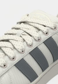 Adidaș de culoare gri deschis cu partea superioară texturată, trei dungi din cauciuc gri închis și șireturi plate cu puncte. Prezintă un vârf rotunjit și un design cu profil jos.