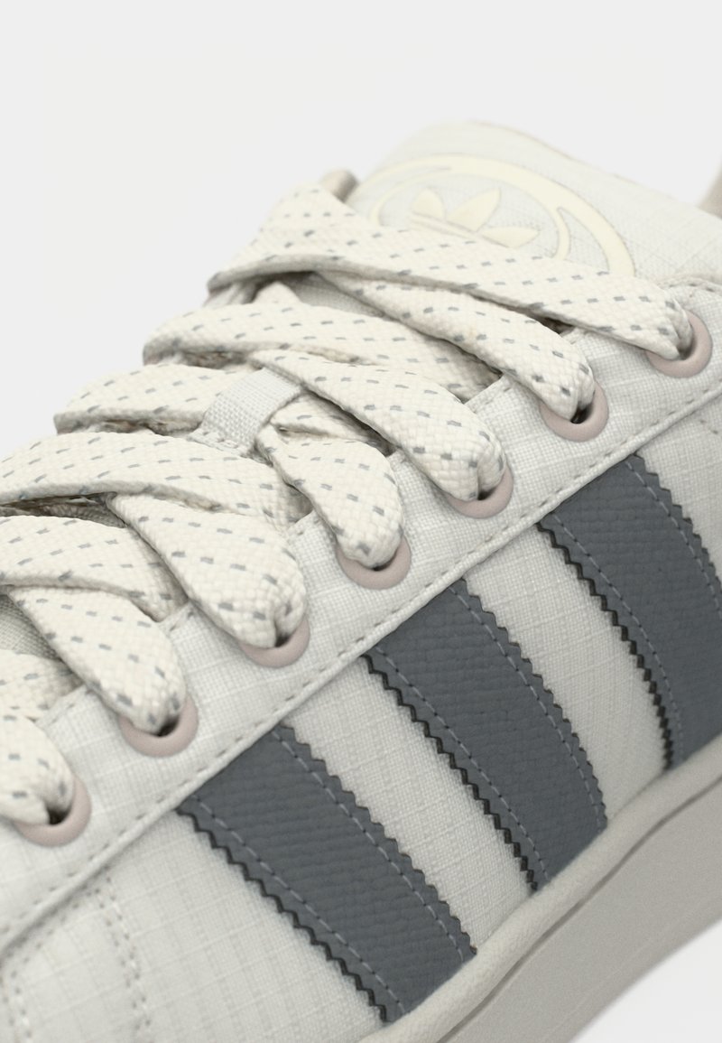 Baskets gris clair avec dessus texturé, trois bandes en caoutchouc gris foncé et lacets plats à pois. Comprend un bout arrondi et un design profil bas.