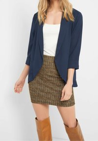Tmavě modrý blejzr s otevřeným předním dílem z lehké látky, spojený s hnědou sukní se strukturou a mřížkovým vzorem, doplněný hnědými kozačkami sahajícími ke kolenům.