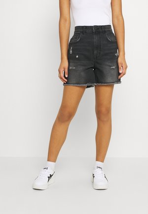 Person iført højtaljede sorte denimshorts med frynset kant og små slidte detaljer, hvide sneakers og en hvid top.